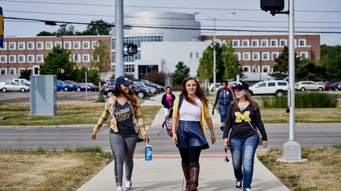 Three young women in UM-Dearborn clothing walk across the street to the Union with the CASL building behind them.