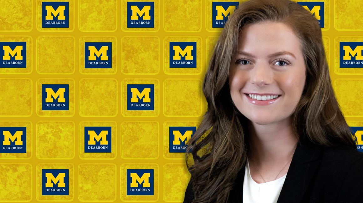 Cady Ericson is a young white woman with green eyes and light brown, shoulder length hair parted on the side. She is smiling against a checkered backdrop that is patterned with the UM-Dearborn logo and maize squares. Cady is wearing a black blazer over a white blouse and a thin silver necklace.