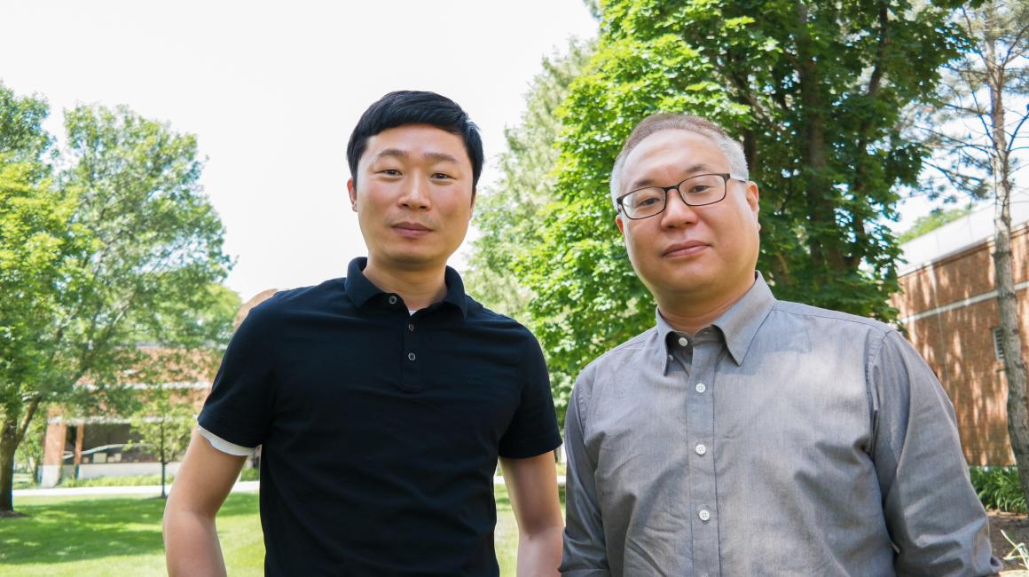 Professor Feng Zhou (L) and Professor Sang-Hwan Kim (R) stand side by side outside behind the ELB. Zhou is a middle-aged Chinese man with short black hair that is swept over. He is wearing a black short-sleeve polo. Kim is a middle-aged Korean man with short gray hair and rectangular glasses. He is wearing a gray, long-sleeve button down.