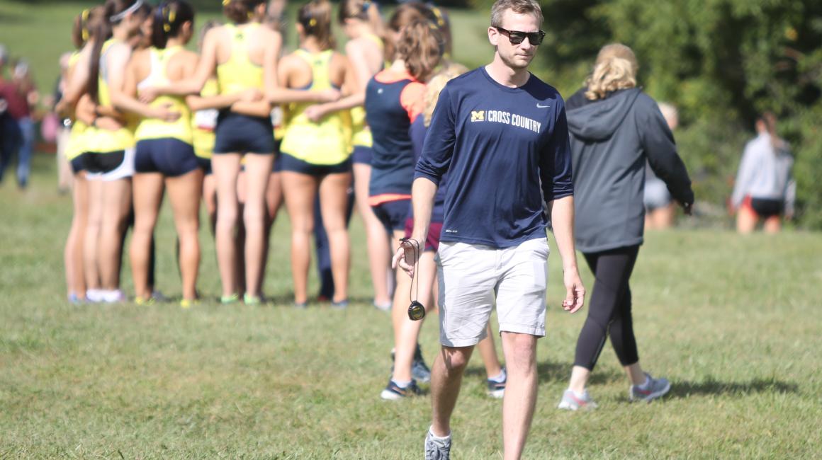 Joe Horka is a middle-aged white man with blond hair. He is walking and wearing a long-sleeve UM-Dearborn Cross Country shirt with the sleeves rolled up and a pair of white shorts. Behind him, in blurry focus, is a group of young women huddled together. They are all wearing blue running shorts and yellow racerback tops with their hair tied up.