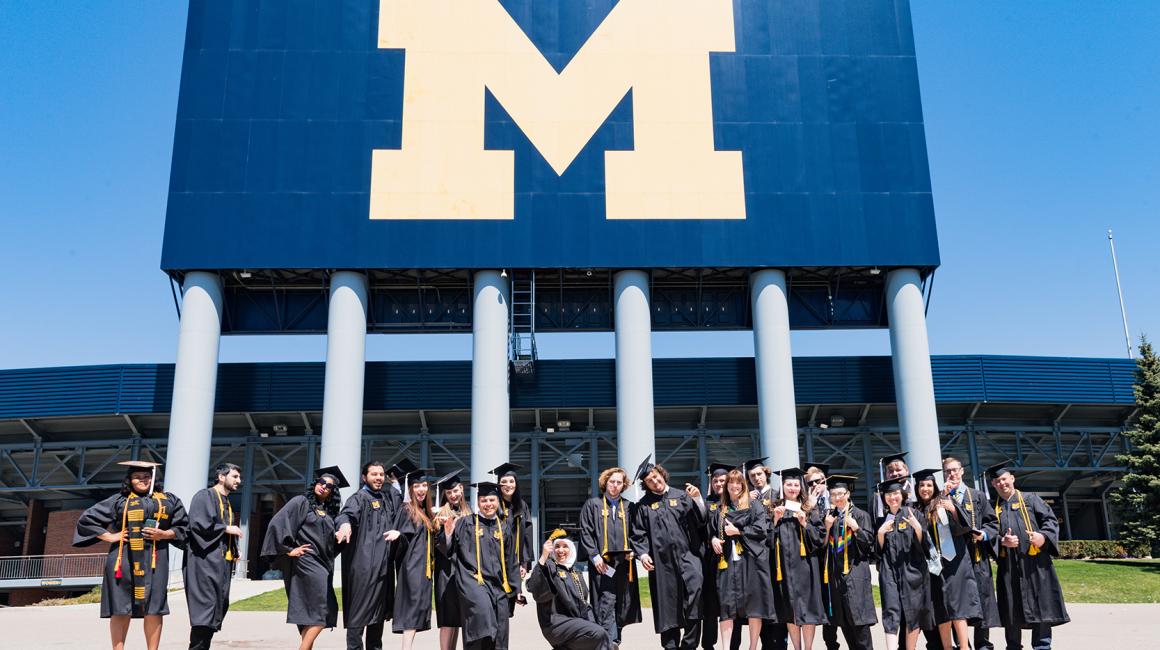 A large group of graduates wearing their caps and gowns from the University of Michigan - Dearborn. They stand in front of the Michigan Stadium.