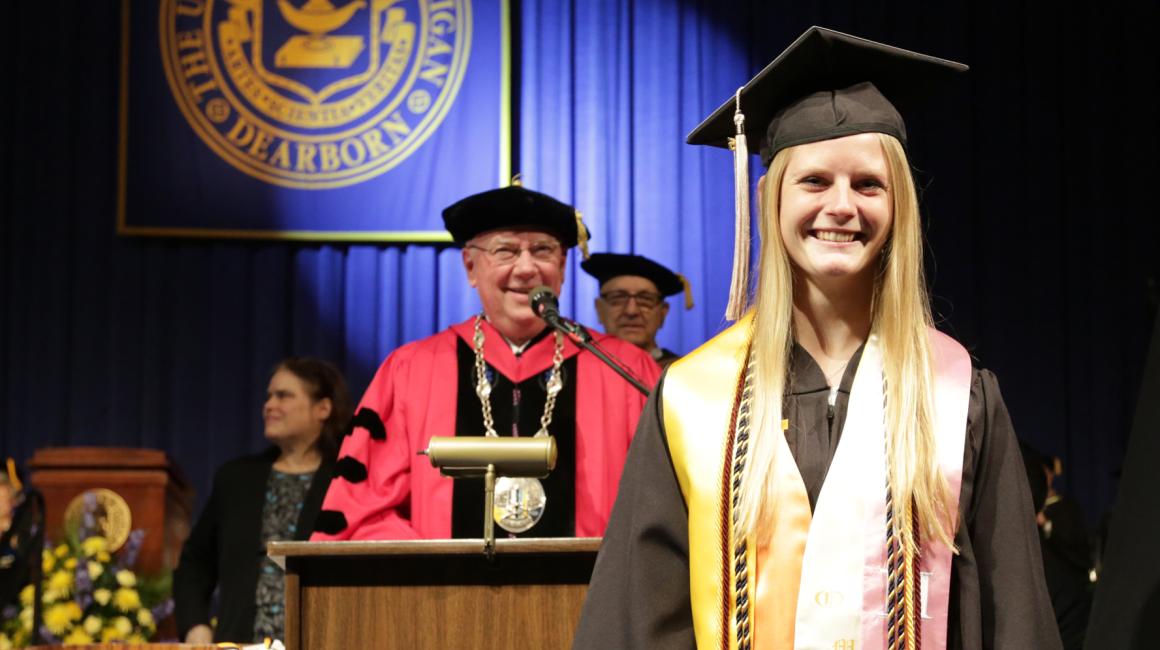 A smiling Megan McDonald stands in front of Chancellor Daniel Little. She is a young, white, blond girl wearing a black cap and gown with a gold and pink stole and four cords. The Chancellor is an older, white male with glasses and is wearing red and black robe and black cap. He is standing on the graduation stage before a podium.