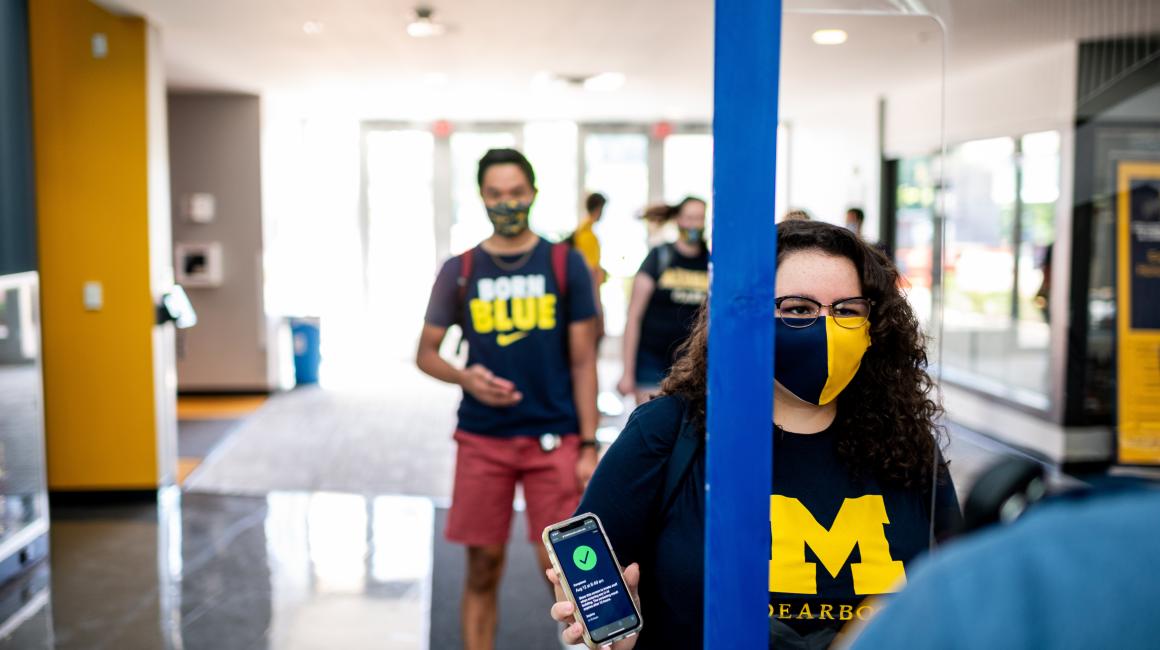 A young, white-presenting woman with black, shoulder-length curly hair and cat eye glasses is lined up at a building entrance holding her phone up. She is wearing a maize and blue “M Dearborn” shirt and split color mask. Her phone shows her health status app with a green check mark icon. Other students (out of focus) stand behind her.