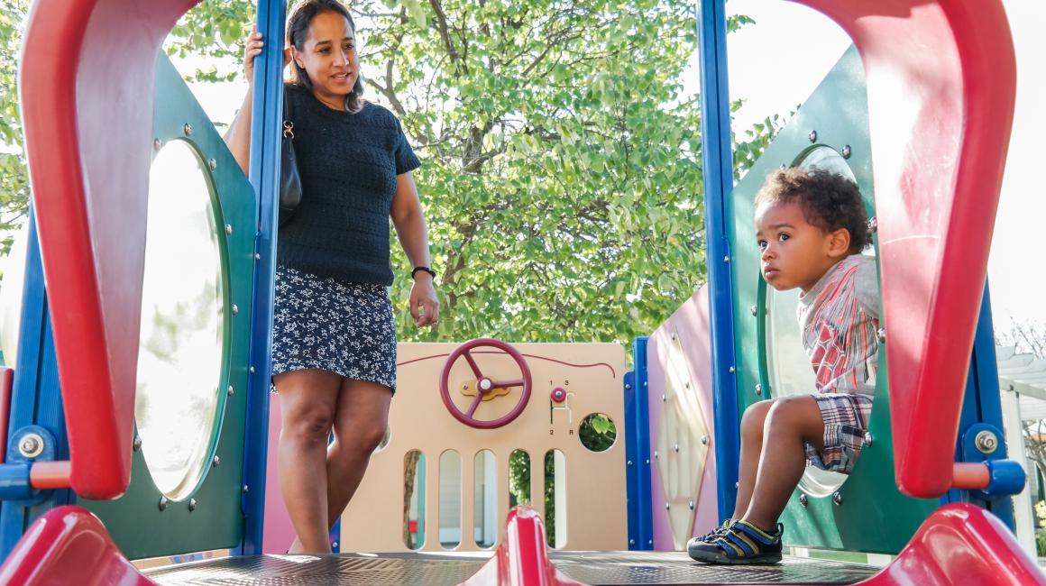 Professor Dara Hill is a middle-aged black woman with straight, brown shoulder-length hair. She is standing in a playground smiling. Dana is wearing a navy blue, short sleeve knit shirt and a navy and white floral patterned skirt just above her knees. Her son Hugh is a toddler with short, curly brown hair and is wearing plaid shorts and a gray and red shirt. He is sitting on a play tunnel entrance.