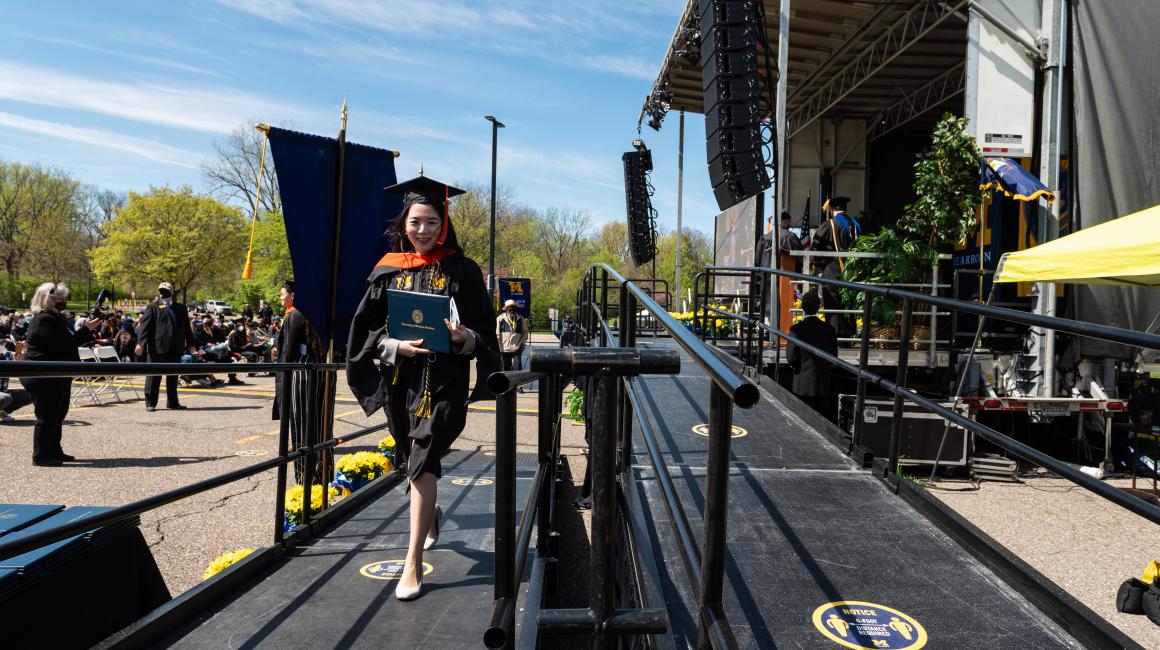 Hyunjoo Park, a Human-Centered Design & Engineering Master's graduate, is a young Korean woman with long, black, straight hair. She is wearing the black UM-Dearborn graduation gown with an orange stole and two maize-and-blue twisted cords with white kitten heels.