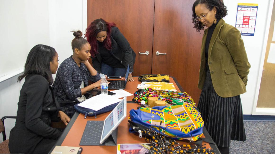Four Black women stand around a table with vibrant, African products.