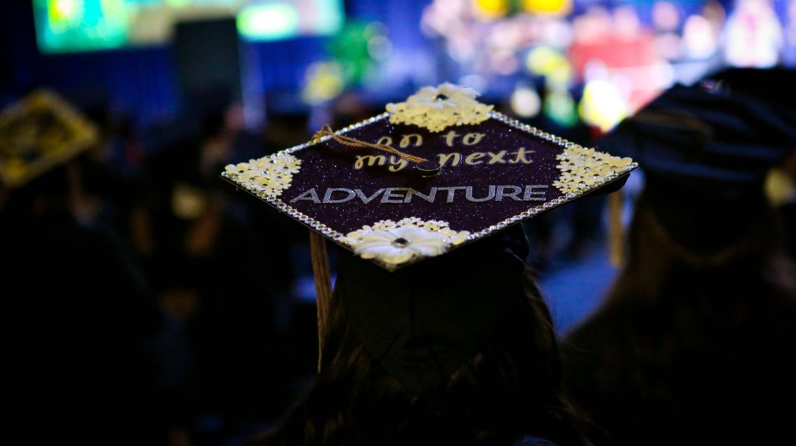 During the graduation ceremony, a shot of the top of a young woman's cap. The cap is decorated with a navy glitter cardstock base, tiny fake white flowers on each corner, and rhinestoned borders. In the center, there is text that reads, "on to my next" in all lowercase, yellow, cursive lettering and underneath in all capital, print, glittery silver lettering "ADVENTURE"