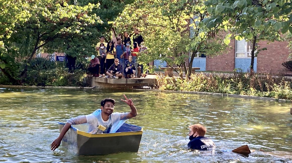 Two students in the Chancellor’s Pond during the 2021 Cardboard Boat Race. One student’s boat has sunk, and the other student’s boat is still afloat. That unsunk student is grinning, holding up a peace sign.