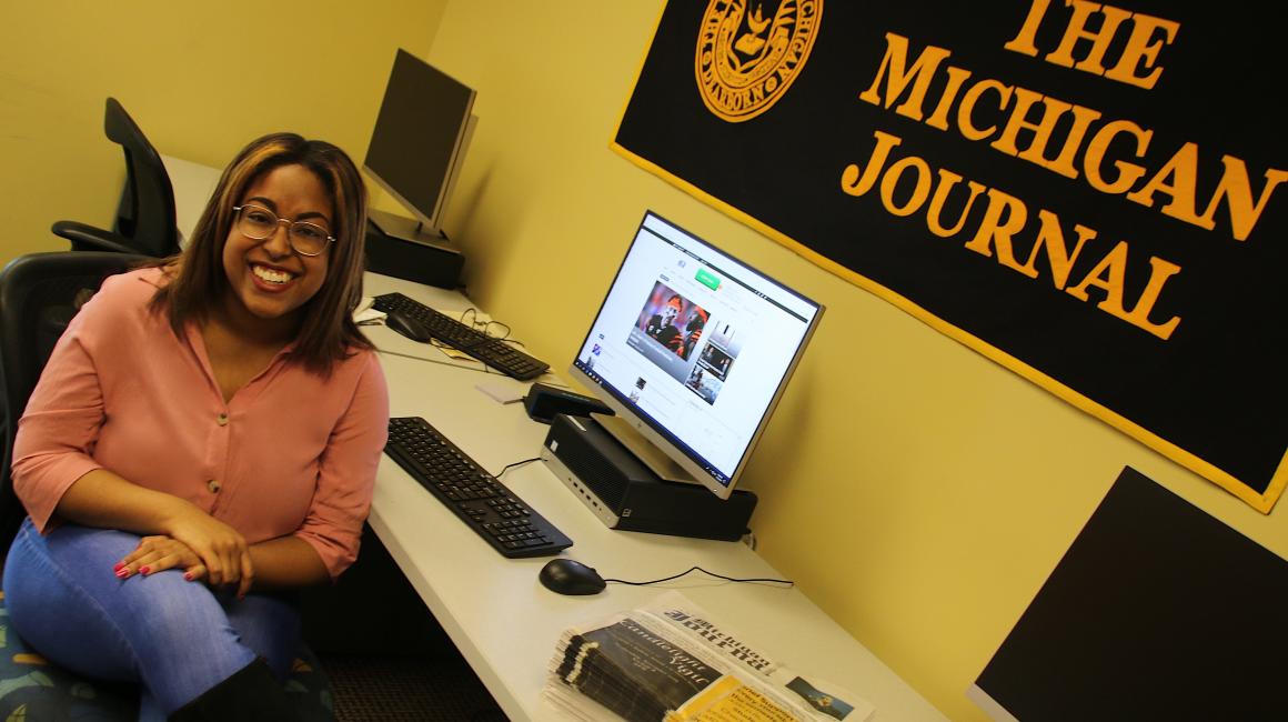Chanel Stitt is a young Black woman with brown/highlighted shoulder-length straight hair and brown eyes. She is sitting at a computer at “The Michigan Journal” with her legs crossed. Chanel is wearing a pair of wire-frame glasses, a pink v-neck button down and a pair of denim skinny jeans.
