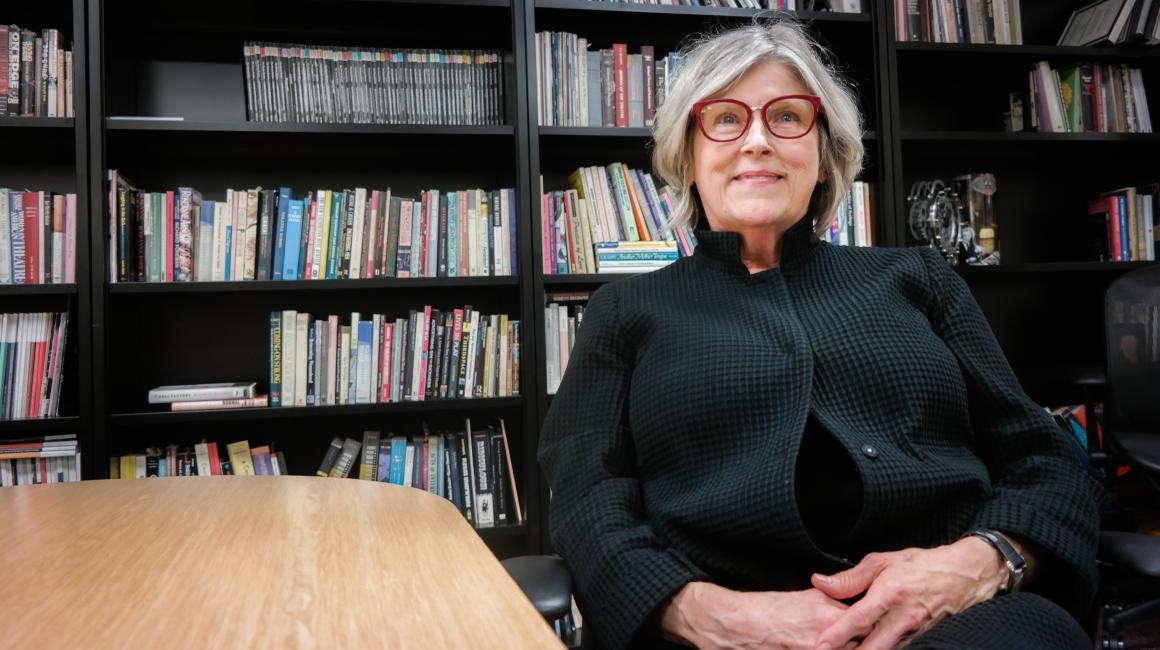 Catherine Davy is an elderly white woman with chin length gray and white hair, blue eyes, and a pair of red, cat eye glasses. She is sitting in front of a bookshelf, wearing a black button cardigan.