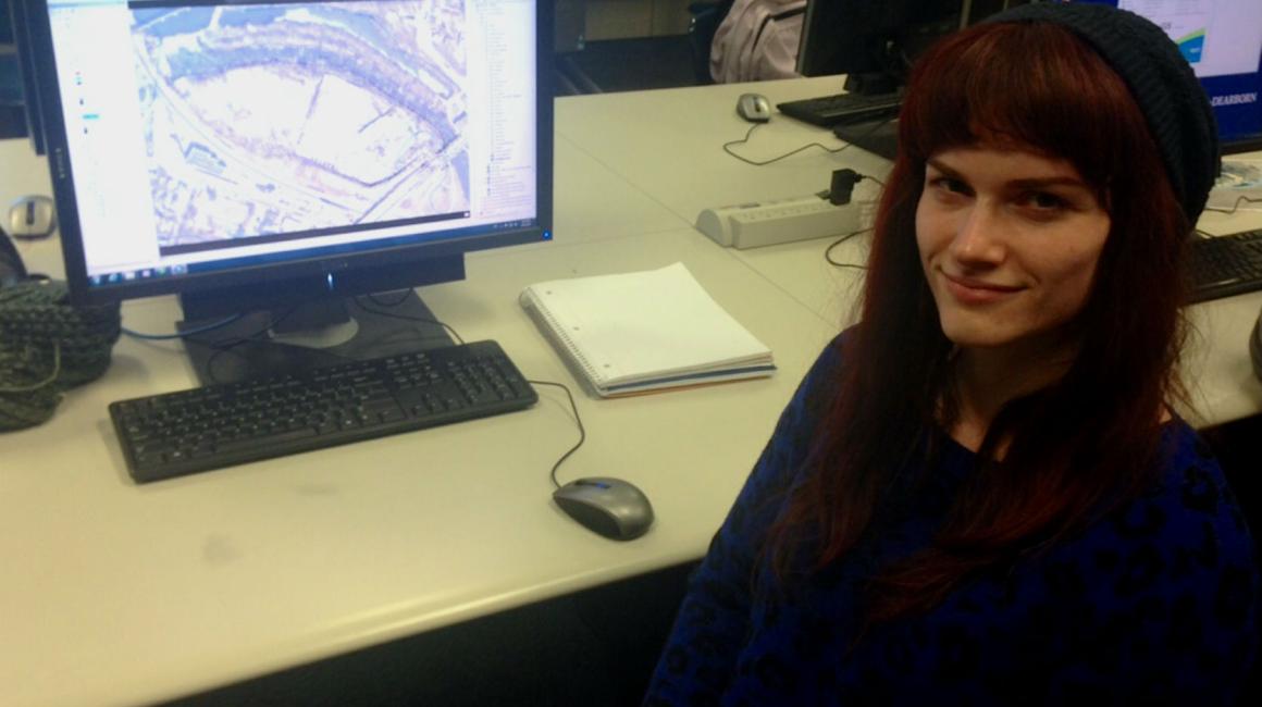 Kelly Williamson is a young white woman with straight, auburn hair that ends past her shoulders and bangs. She is wearing a navy sweater and black beanie. Kelly sits in front of a computer with the GIS open.