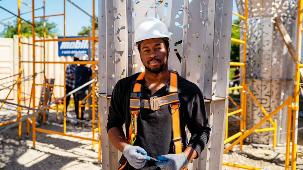 Keviyan Richardson is a Black man with short black hair and facial hair. He stands in front of scaffolding in a construction site. He is wearing a black shirt with rolled up sleeves, an orange safety harness, a white safety helmet, and gray and blue gloves.