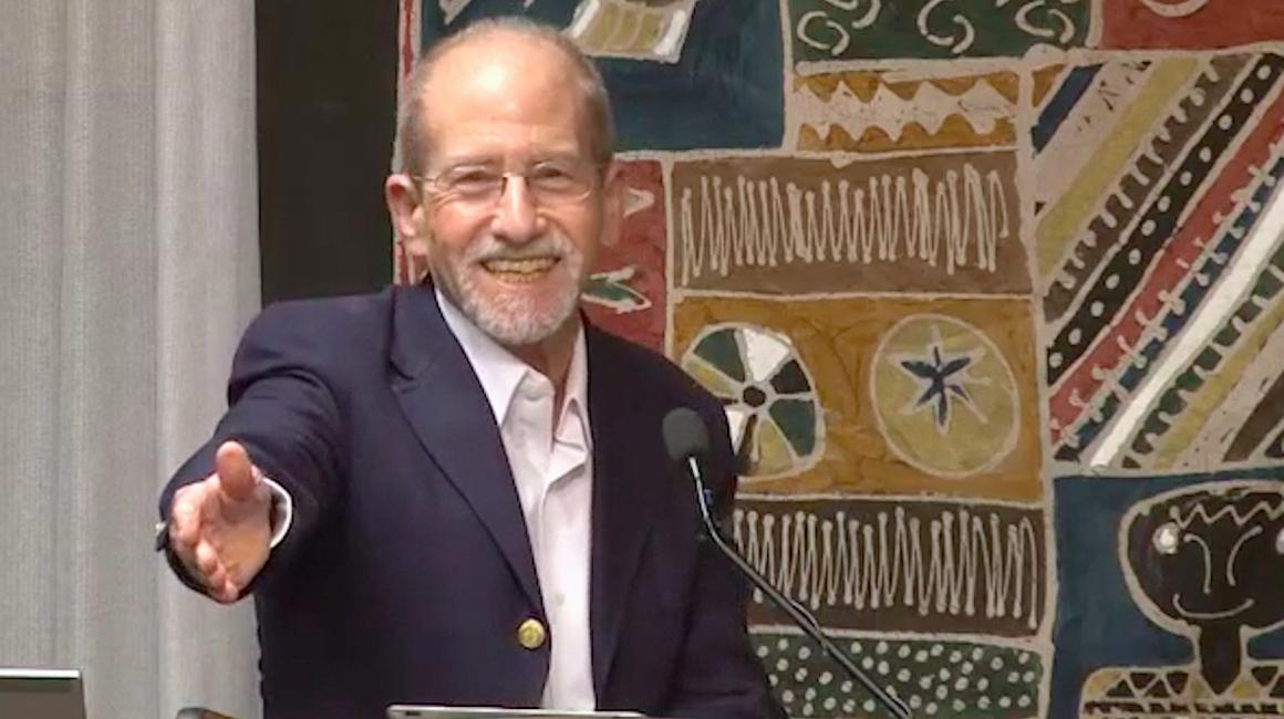 Elias Baumgarten is an older, white man with gray, thinning hair and facial hair. He is wearing a navy suit jacket with a white button down and thin, wire frame glasses. He is standing behind a podium smiling and holding his hand out.