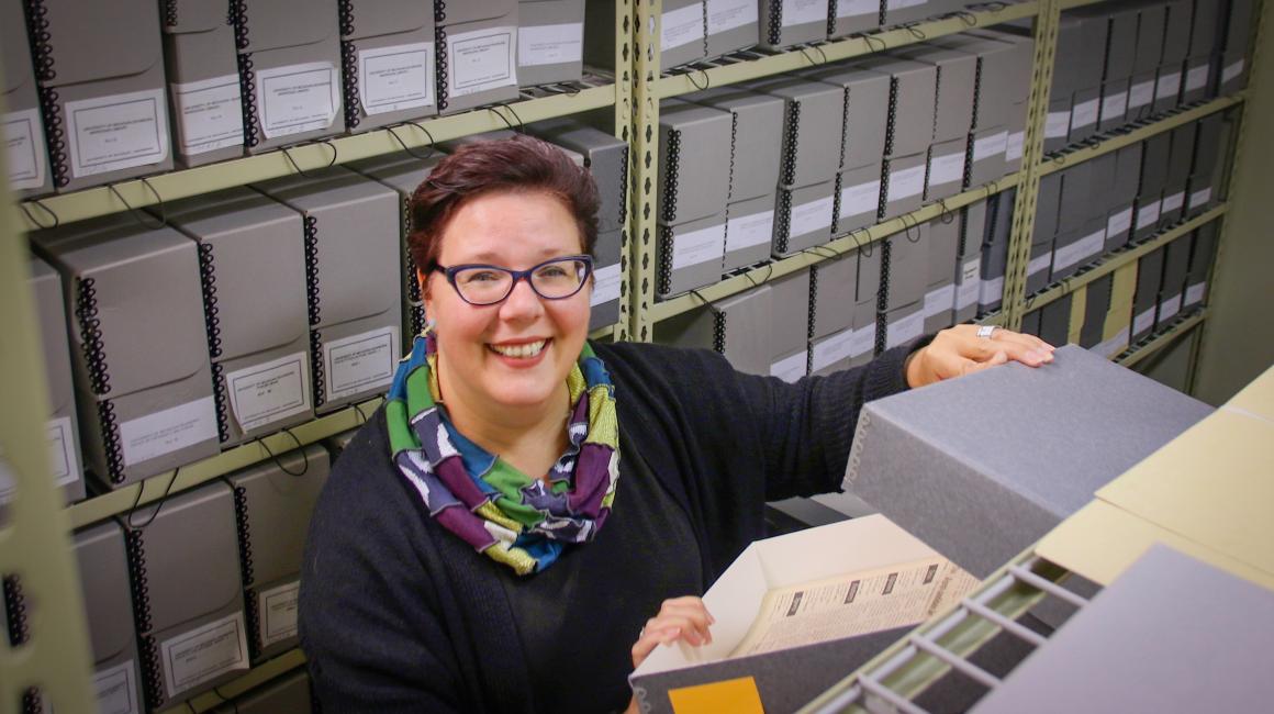 Julia Banel Walkuski is a middle-aged white woman with short, auburn-dyed hair. She is wearing a pair of purple cat eye glasses, a black sweater, and patched infinity scarf. Julia is sitting in an archive room and is opening an archive box.