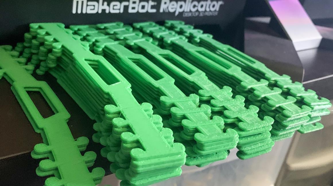 About 60 bright green 3D printed face mask extender straps sit on the bed of a 3D printer.