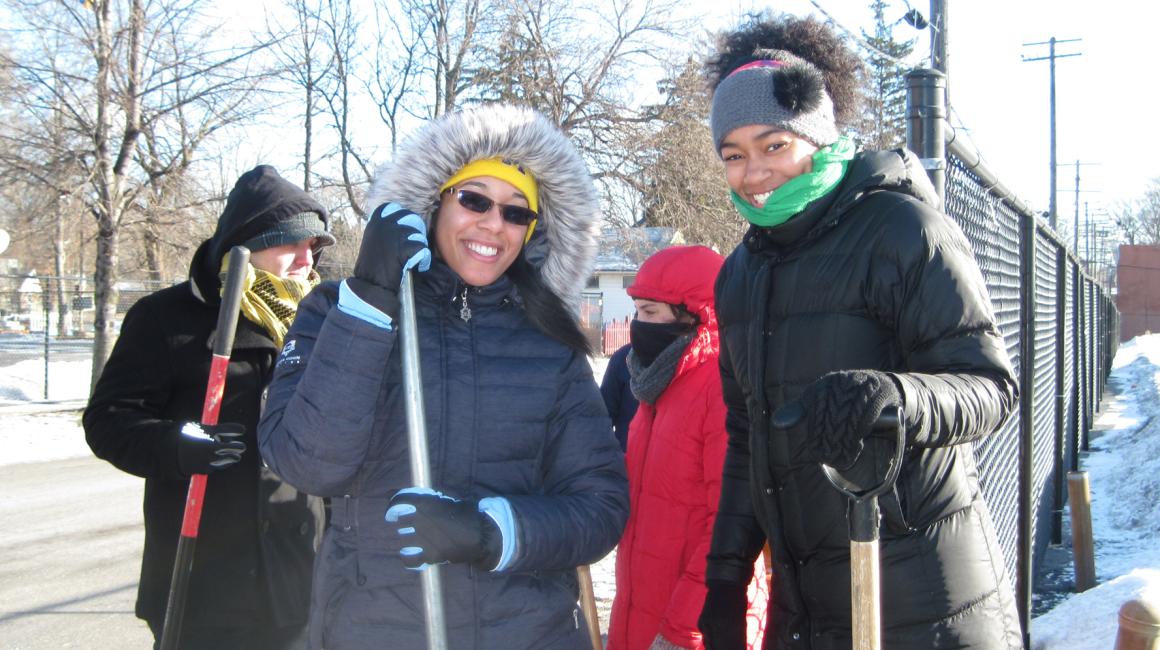 Four students stand outside in the snow holding shovels and wearing thick puffer jackets.