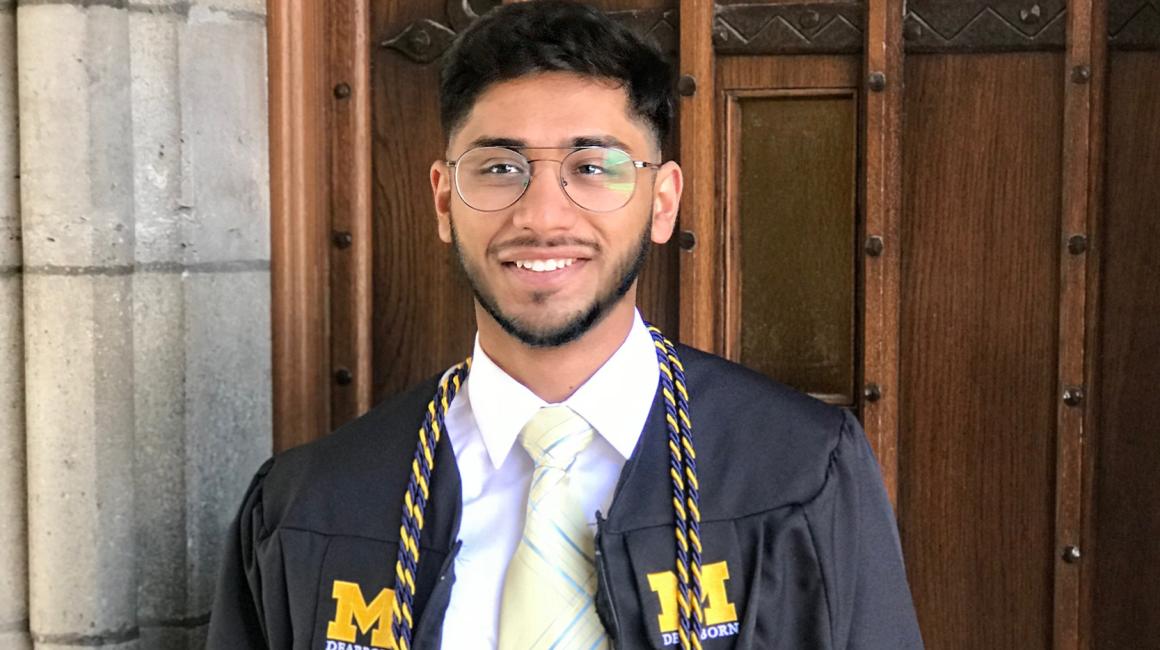 Muhammad Osto is a young Middle-Eastern man with black hair and facial hair. He is wearing oval aviator style glasses, a white button up with a yellow and blue striped tie, and a UM-Dearborn graduation gown with two maize and blue striped cords.