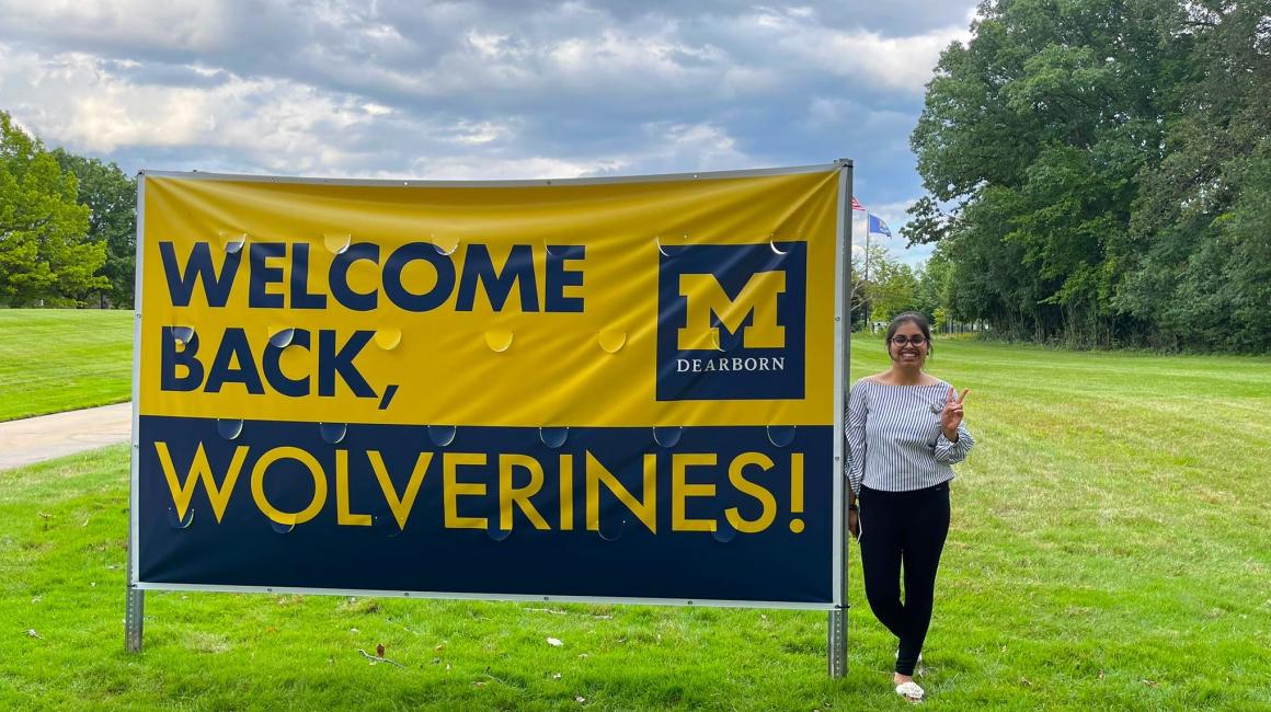 Richard Chachra is a young South-Asian woman with dark brown hair tied back in a bun and loose bangs and black rectangular glasses. She is wearing a black-and-white vertically striped blouse and black leggings. She is standing and holding up a peace sign next to a maize and blue vinyl poster that says “WELCOME BACK, WOLVERINES!”
