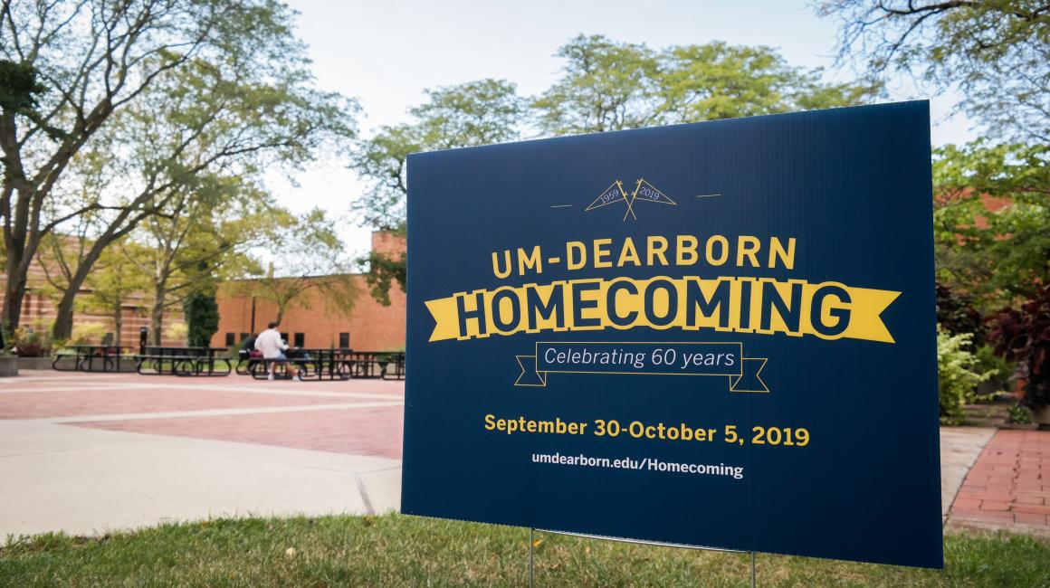 A navy blue poster staked out in the common area around the Chancellor’s Pond. On the top of the poster, there are two outlined flags reading “1959” and “2019”. Underneath, it reads in all capital, yellow text: “UM-Dearborn Homecoming”, “Celebrating 60 years”, “September 30 - October 5, 2019”, “umdearborn.edu/Homecoming”
