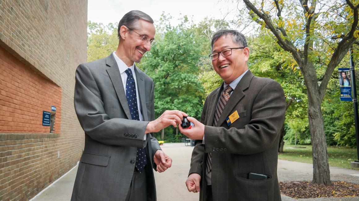 Ed Krause and Taehyun Shim stand side by side, holding a control system part. Ed (left) is a middle-aged white man with slicked back gray hair and goatee. He is wearing a pair of rectangular frameless glasses, a white button up with a navy-and-white polka dot tie, and a gray suit jacket. Taehyun (right) is a middle-aged Korean man with gray hair and no facial hair. He is wearing a pair of black rectangular glasses, a blue button up with a brown-and-white striped tie, and a brown suit jacket.