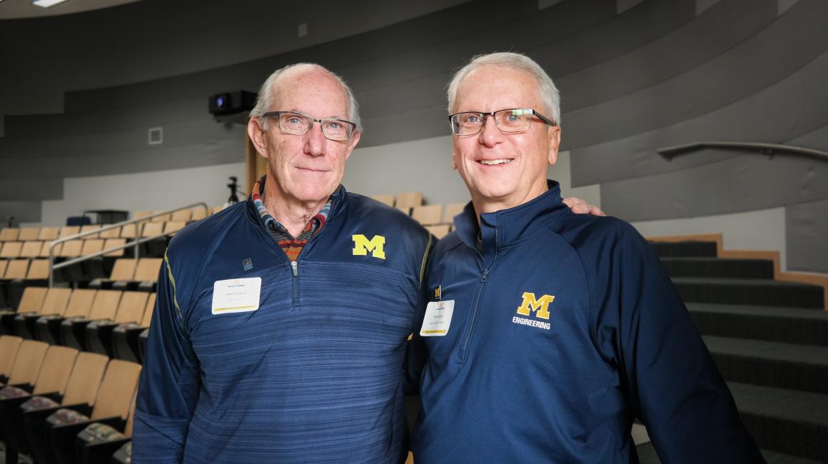 Jim Gingrich (L) and Tom Demmon (R) are two elderly white men. Jim has thinning gray hair and green eyes. Tom has thicker gray hair and green eyes. Both are standing side-by-side with arms hooked on one another's shoulders and are wearing UM branded quarter-zips.