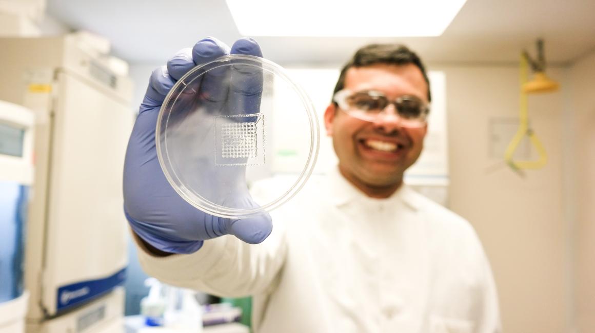 Nilay Chakraborty is a South Asian man with short brown hair and no facial hair. He smiling in the background wearing a white lab coat and blue gloves. He is blurred out of focus and is holding up a Petri dish with a clear square of bio 3D printed material.