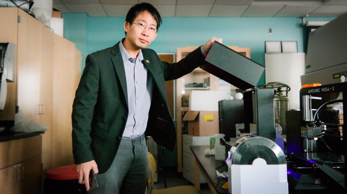 Professor Jo is a middle-aged Chinese man with black side-parted hair and thin rectangular wire glasses. He is wearing a light purple button down tucked into a pair of gray denim and a black blazer over top. He is facing the camera and standing next to a 3D printer.