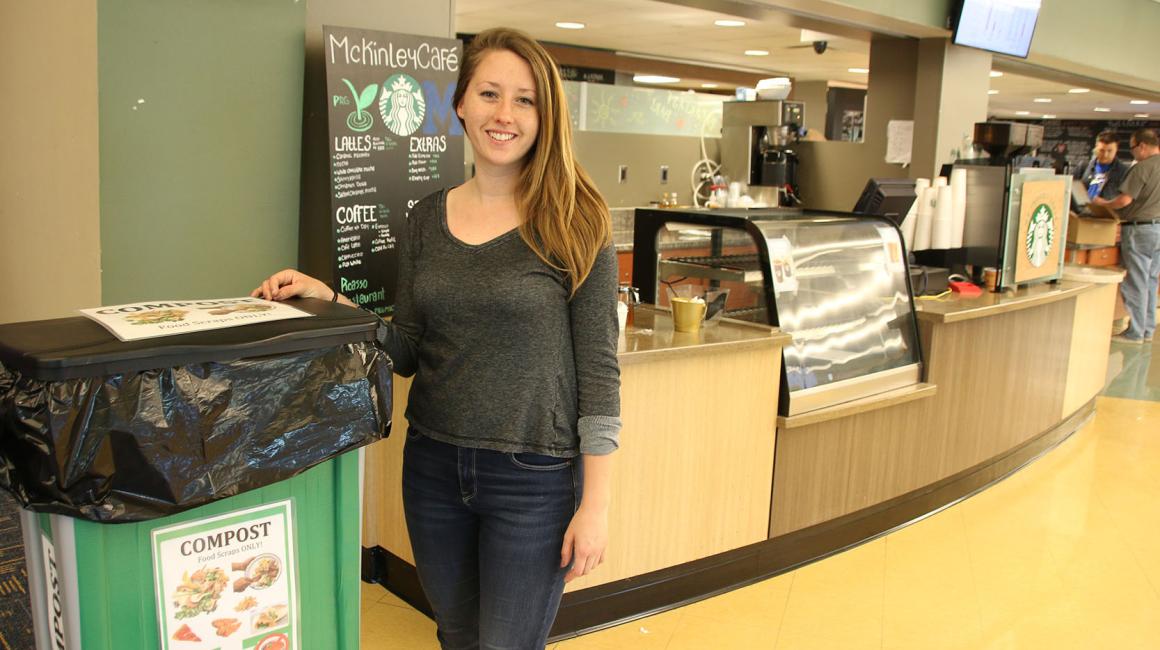 Sarah Bellaire is a young, white woman with straight, dark blonde hair that lays past her shoulders. She is wearing a dark, heather gray long-sleeve shirt and a pair of dark denim skinny jeans. She stands next to a green compost bin inside of the McKinley Cafe.