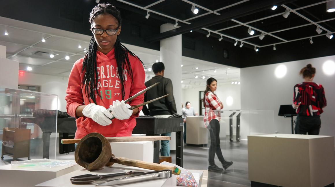 Kari Perkins is a young Black woman with chest-length braids draped over her shoulders. She is wearing a pair of black, rectangular glasses, a red “American Eagle” hoodie, and white gloves. She is handling an ancient artifact with a pair of forceps.