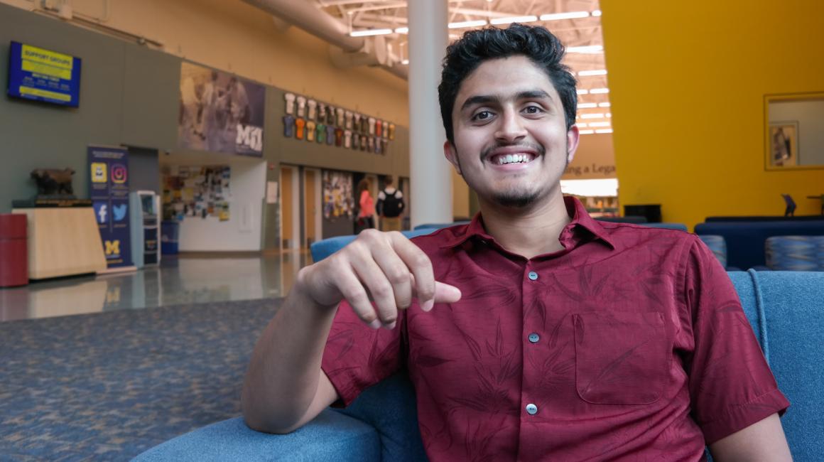 Syed Akbar is a young South-Asian man with swept over black hair and facial hair. He smiling at the camera, sitting in a sofa chair inside the University Center Commons. Syed is wearing a red, short-sleeve button down.