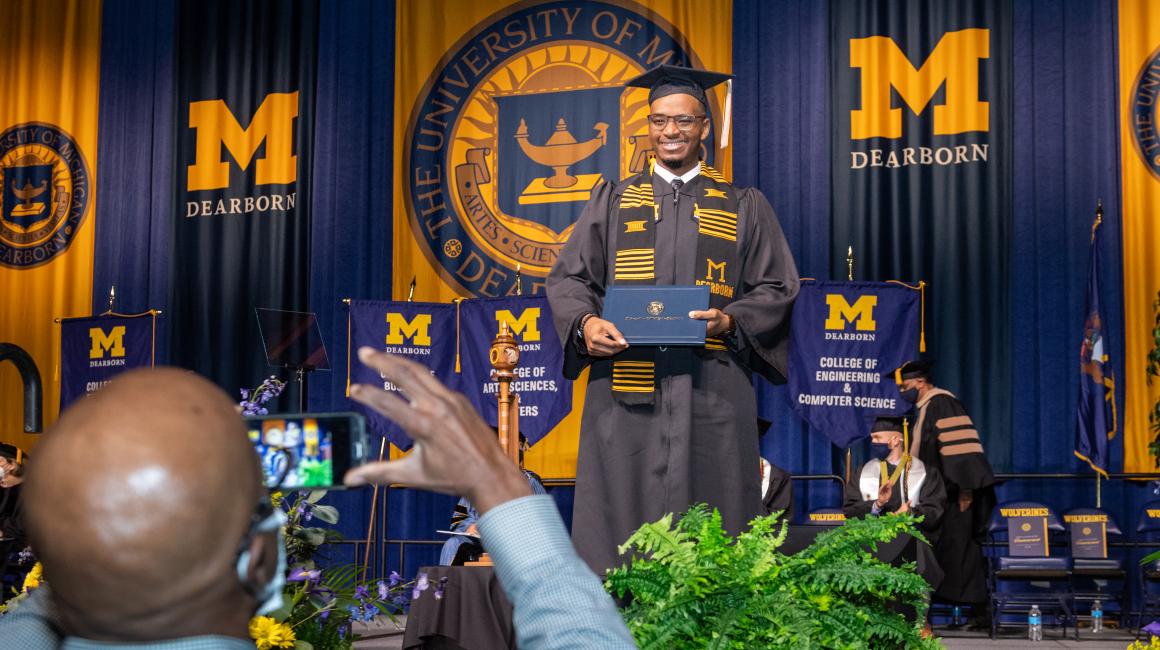 A young Black man wearing his cap and gown, stands for a photo while holding his degree.