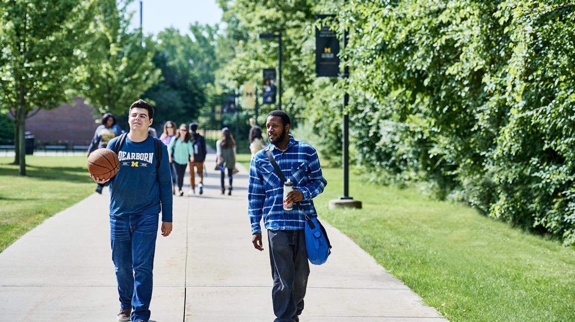 A photo of students walking on the walkway behind the University Center. In focus are two students: the first student is a young white man with short black hair. He is holding a basketball. The second student is a young Black man with short black waves. He is holding a backpack and a white sports bottle. Both are wearing dark blue shirts.