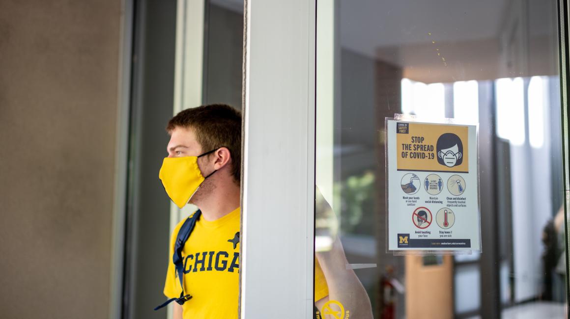 A young white man with short strawberry blond hair is walking out of the SLRC doors. He is wearing a bright maize crew neck short sleeve and a maize-and-blue split color mask. A informational graphic sign on the door reads “Stop the spread of COVID-19”