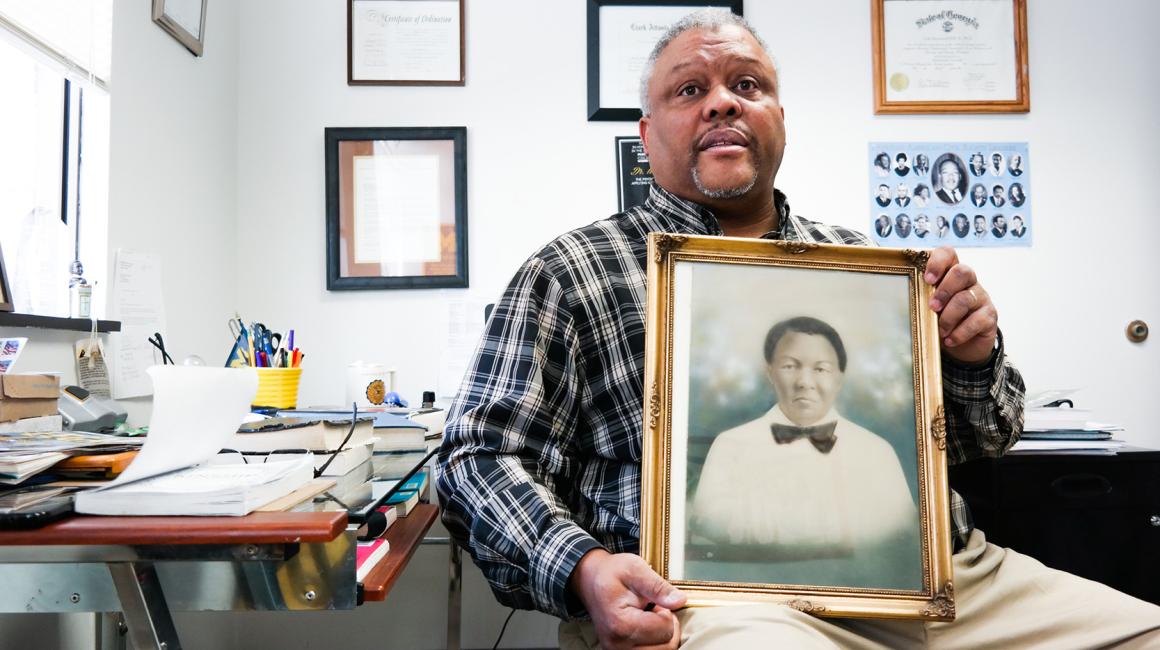 Ural Hill is a middle-aged Black man with gray hair and a goatee. He is wearing a black-and-white plaid shirt, holding a portrait of his great-great-grandmother, Rachel Stewart, a young Black woman with short black hair.