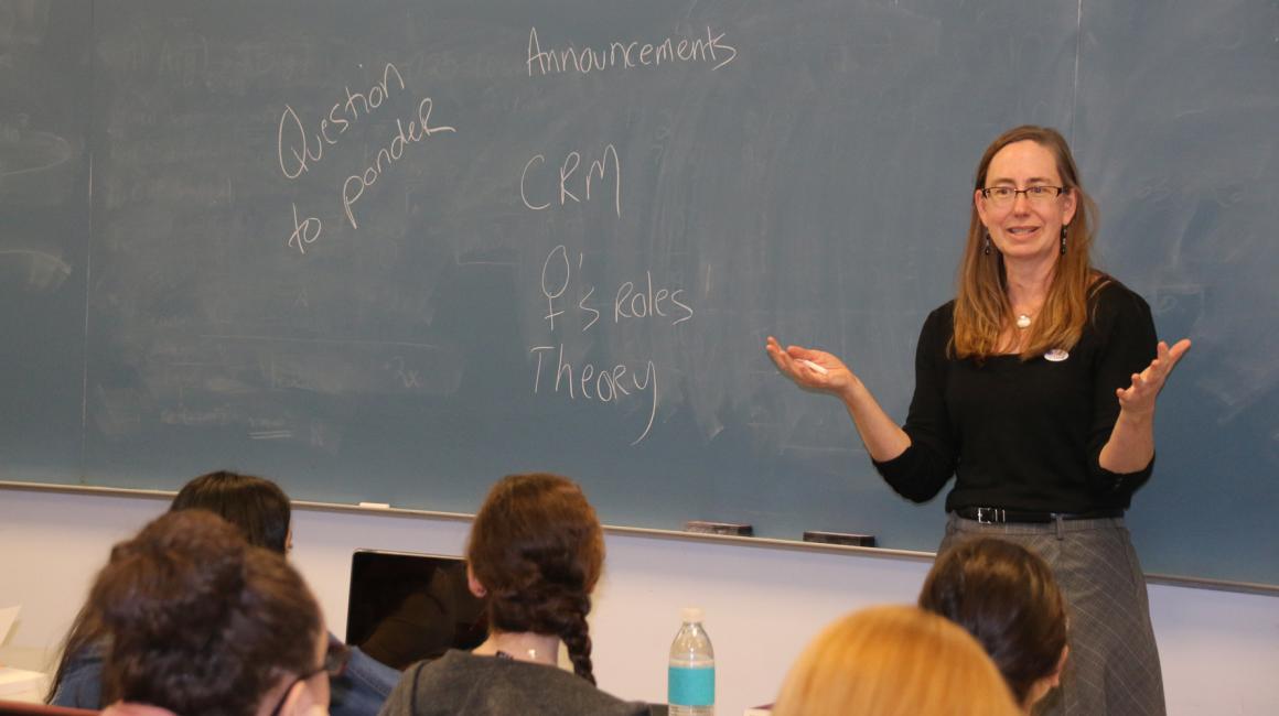 Professor Georgina Hickey stands in front of a blue-gray chalkboard with her hands out in mid-explanation. Written on the chalkboard: “Question to Ponder”, “CRM, Women’s Roles, Theory”, “Announcements” Georgina is a middle-aged white woman with straight, dirty blonde hair laying past her shoulders. She is wearing a black, square neck long sleeve shirt tucked into a pair of plaid trousers.