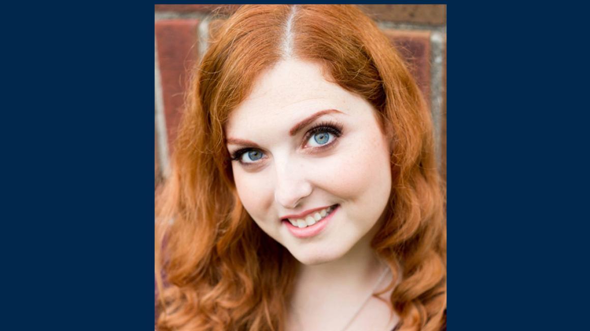Adriane Galea is a young white woman with red hair and blue eyes. She is smiling and wearing a deep purple v-neck with a silver necklace.