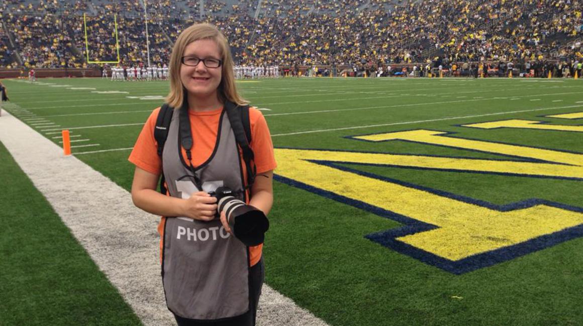 Amber Ainsworth is a young white woman with shoulder-length, straight blonde hair. She wears a pair of black, rectangular glasses, a black backpack, and an orange t-shirt under a gray UMich “Photo” vest. Amber is holding a camera outside of the Michigan Stadium’s end zone.
