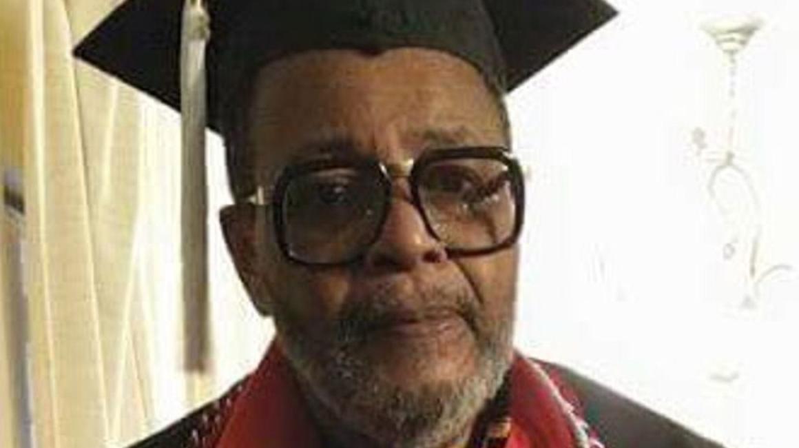 Carlton McPhail Sr. is a senior. Black man with dark hair and a salt-and-pepper beard. He is wearing a pair of thick, square glasses, the UM-Dearborn cap and gown, and two red/white/blue cords, one silver cord, one red cord, & a red stole.