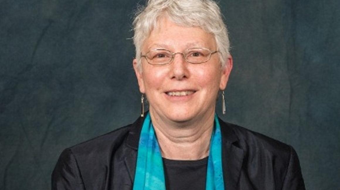 Jacqueline Vansant is an old white woman with short white curls. She is wearing a black cardigan over a black undershirt with a bright teal accent scarf. She is holding the Distinguished Research Award; which is on a walnut plaque with a blue front.
