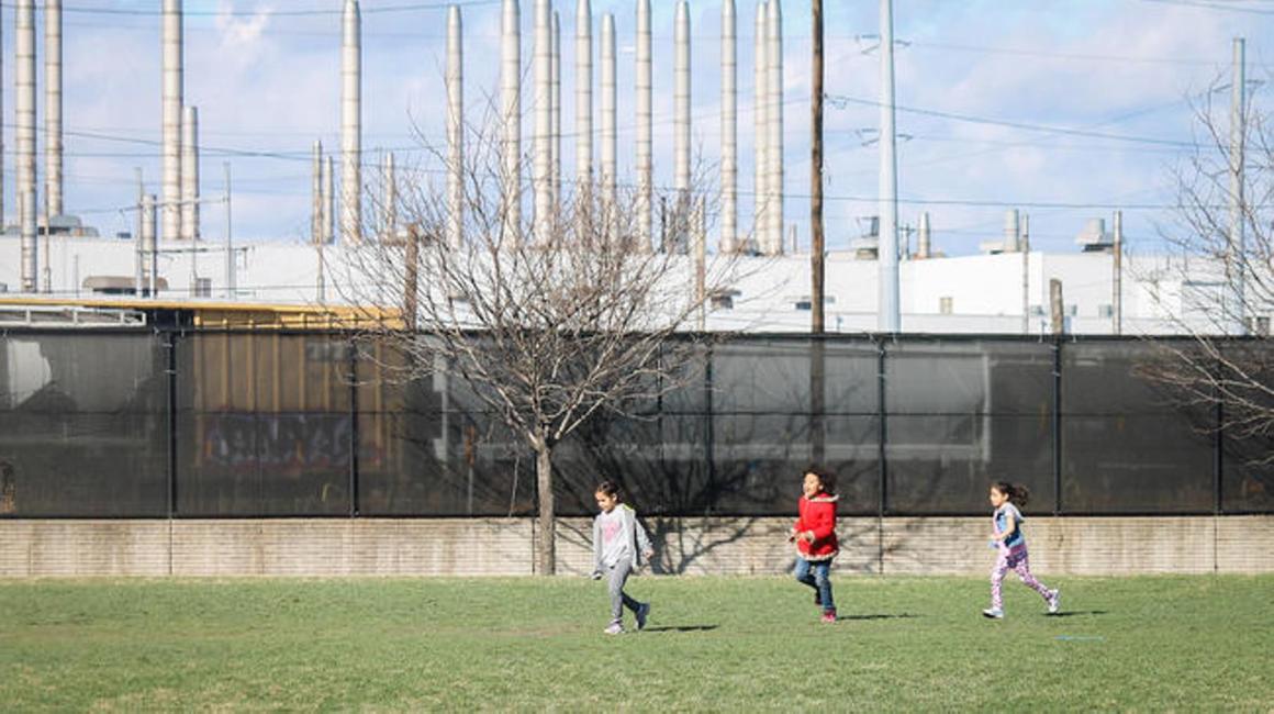 Three children are running and playing in front of a black fence bordering a factory plant.