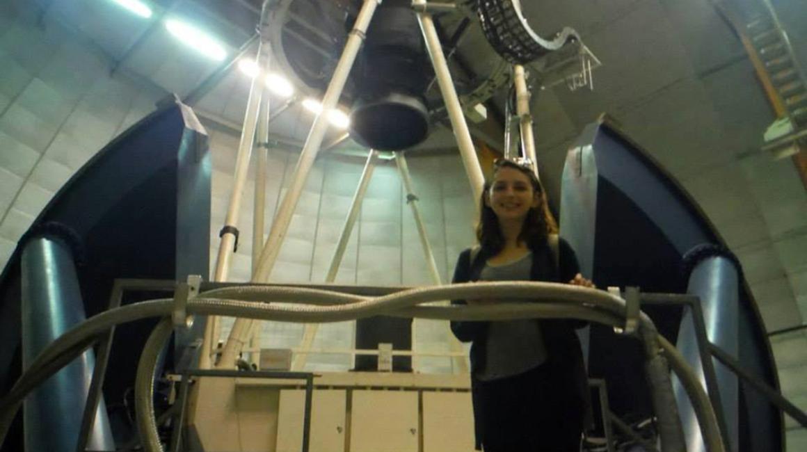 Brittany Howard is a young white woman with shoulder length brown hair. She is standing in an observatory and is wearing a gray tank underneath a black cardigan. Her glasses are set over her head.