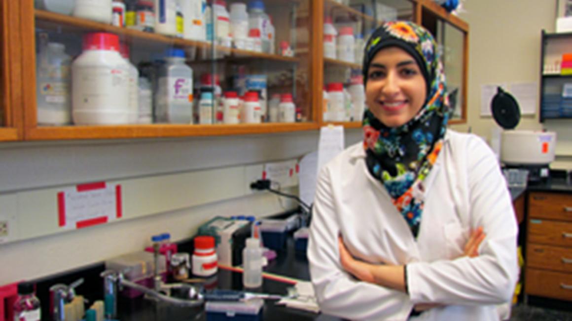 Walaa Tout is a young Arab Muslim woman who is wearing a black hijab with a floral pattern and a white lab coat. She is standing in a lab with various bottles and is smiling while facing the camera and having her arms crossed.