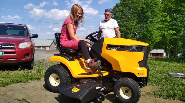 A young woman in a dress and work boots sits on a riding lawmower, flanked by an older man who is explaining how to use it.