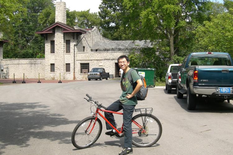 Xuan Zhou poses for a photo on his bike, in front of a historic stone building on the UM-Dearborn campus