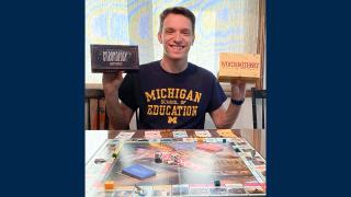 A photo of student Ezra Houghtby, sitting at a table in front of a Monopoly board while holding up boxes of two of his favorite board games.