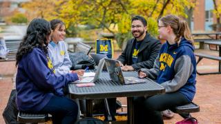 4 students wearing UM-Dearborn clothing site around an outdoor table.