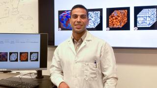 Wearing a white lab coat, doctoral student Hossein Abbasi poses for a portrait in front of computer screens in a lab