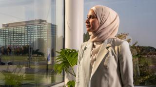 Wearing all white, alum Zaineb Hussein looks out through the large glass windows in her office, overlooking Dearborn, with the Ford corporate headquarters building in the background