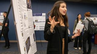 Standing in front of her poster, a student gives a presentation on her research at the Summer Undergraduate Research Experience showcase