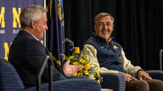 Vice Chancellor for External Relations Ken Kettenbeil talks with Domenico Grasso on stage with a UM-Dearborn banner in the background and a maize and blue flower arrangement between them.